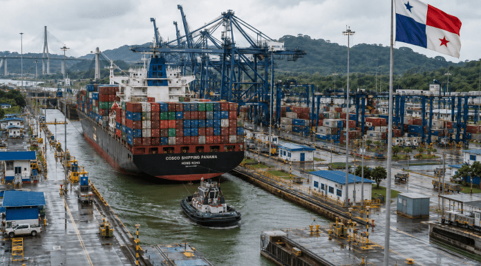 The LAC-Panama Stalemate Continues Container ship transiting a rain-soaked Panama Canal port beneath towering cranes, with the Panamanian flag flying above the terminal.