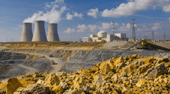 Hyper-realistic landscape showing uranium ore and an open-pit mine in the foreground, with mining equipment, processing facilities, a nuclear power plant with cooling towers, and electrical transmission lines in the background, representing the relationship between uranium term contracts, utility procurement, supply, and spot prices.